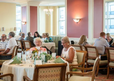 Diverse group of elderly people dining in a brightly lit room with elegant decor, some reading menus.