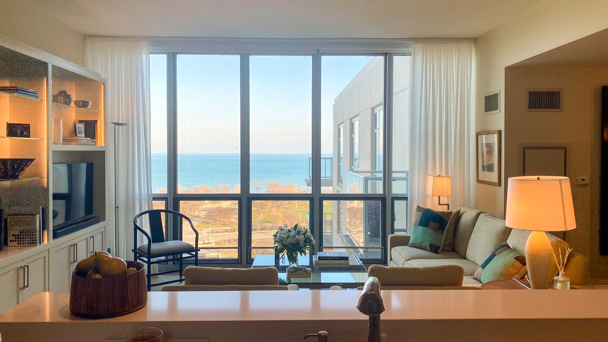 IMG-2407-scaled Interior view of a living room with large window overlooking the sea, featuring a couch, chairs, and shelved decor.